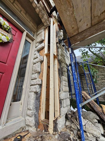 A red door next to a stone-clad wall undergoing repair with exposed wooden studs and construction scaffolding.