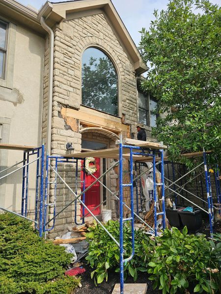 A worker on scaffolding repairs the stone facade and entryway of a two-story home with a bright red front door.