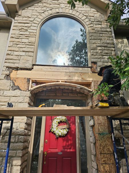A worker on scaffolding repairs the stone masonry and wood framing around a red front door beneath a large arched window.