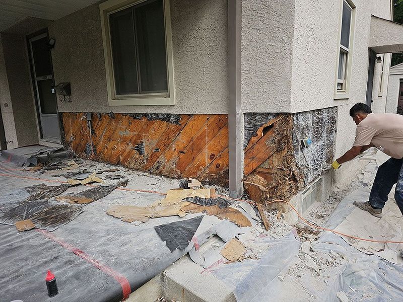 A worker repairing a damaged exterior wall on the lower section of a building, removing old siding and exposing wood.