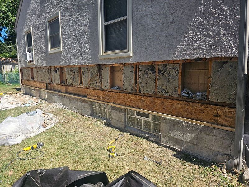 The exterior wall of a house has been stripped of siding, exposing the wood sheathing and insulation above the foundation.