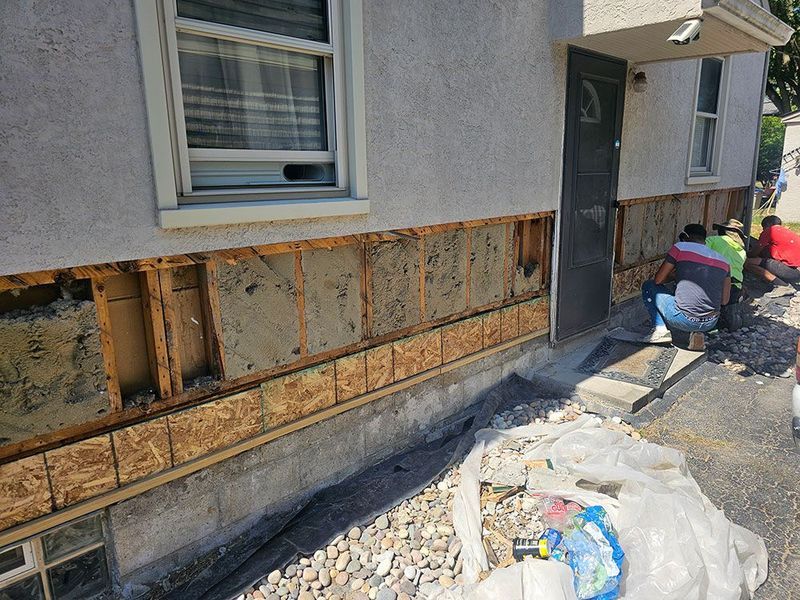 Workers replace the exterior siding of a house, revealing the wooden frame and insulation beneath the wall.
