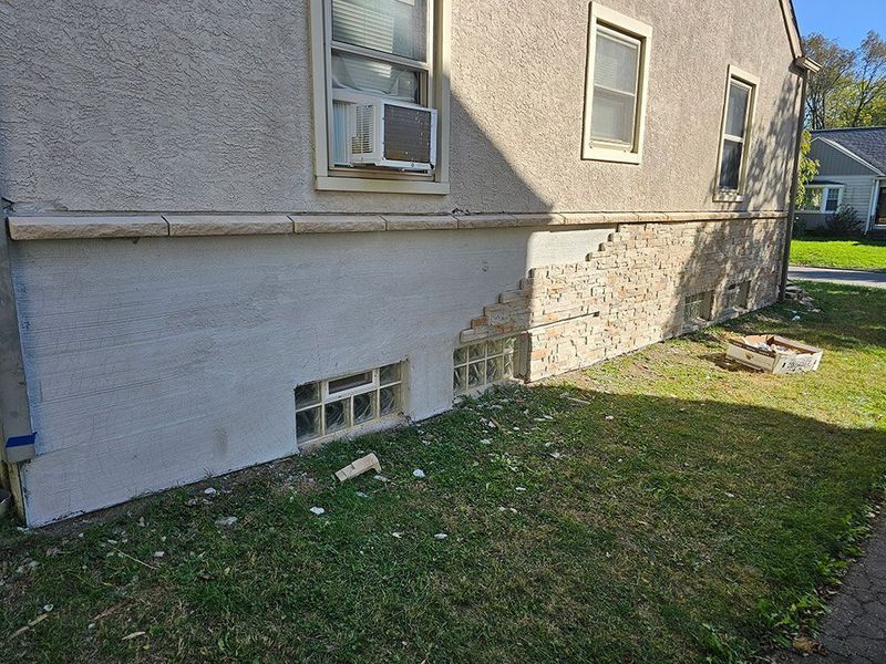 Side view of a house foundation showing unfinished repairs, with half the stone wall covered in fresh light-gray plaster.