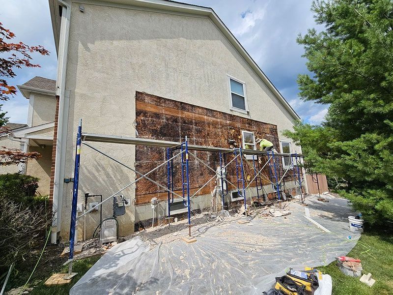 A construction worker on scaffolding repairs a house exterior where stucco has been removed to expose the wooden framing.