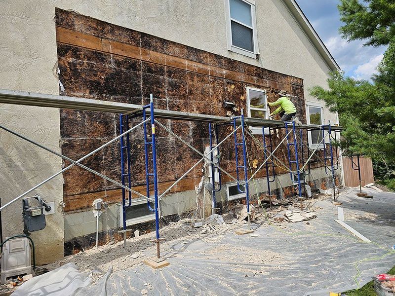 A construction worker on scaffolding repairs the exterior wall of a house, where the stucco has been removed.
