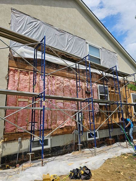 A house under renovation with exterior siding removed, revealing wood framing and pink insulation, with blue scaffolding.