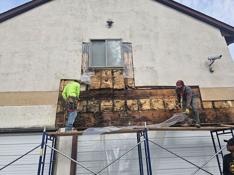 Two workers on scaffolding repair a section of rotted wood sheathing beneath a second-story window on a stucco house.