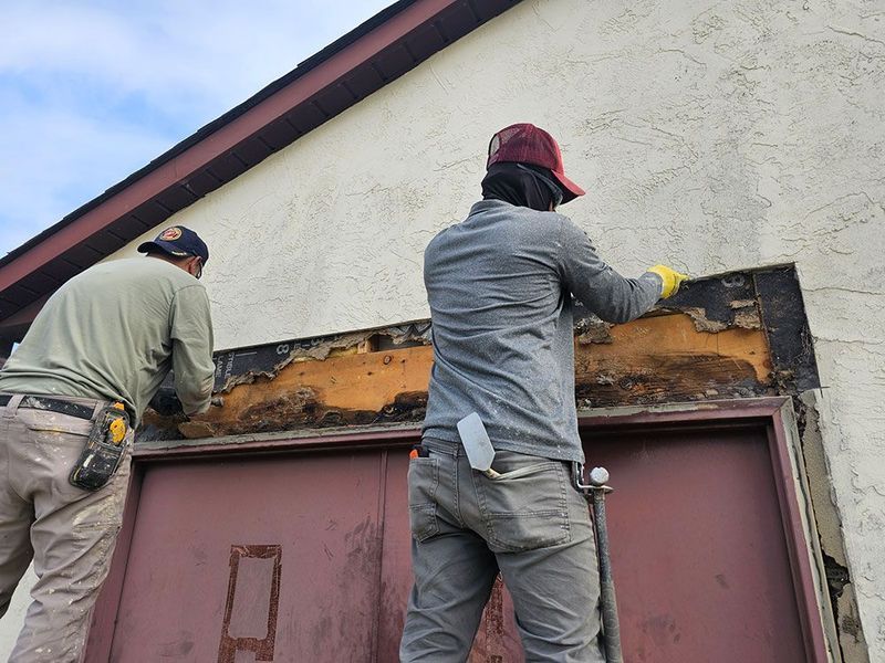 Two people in work clothes repair the area above a maroon double door on an exterior building wall.
