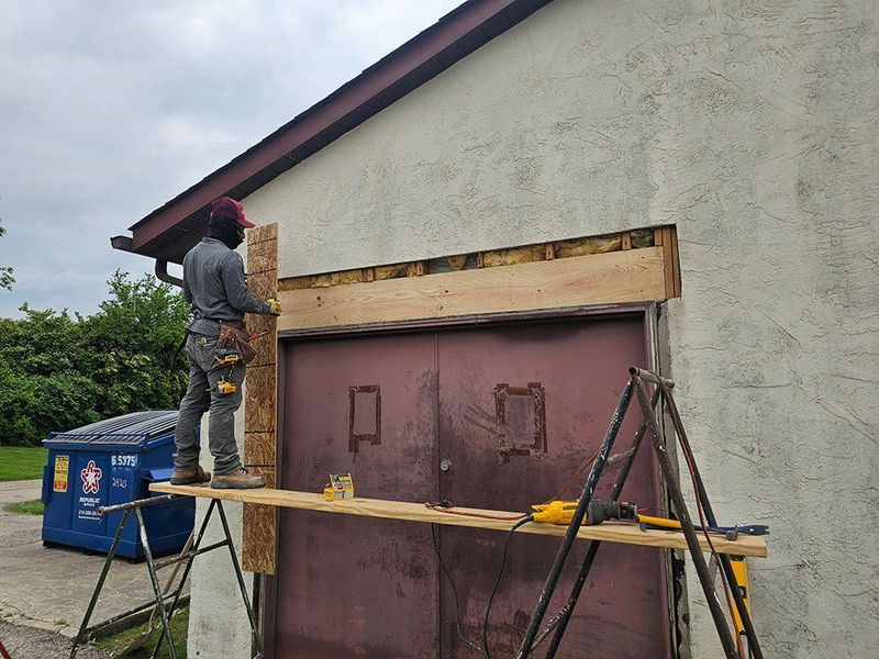 A construction worker stands on a scaffold while installing a wooden header above a doorway on a stucco building exterior.