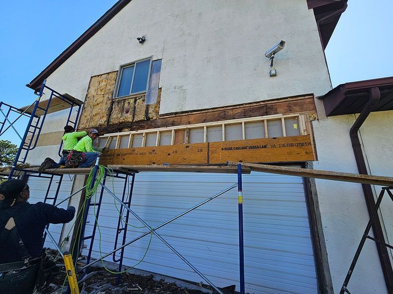 Construction workers on scaffolding repair the exterior wall above a garage door, installing new wooden framing.