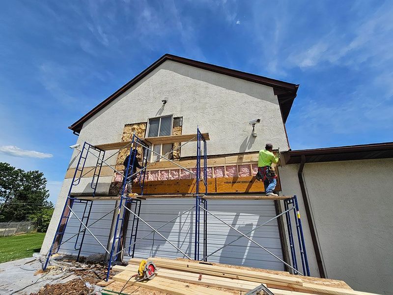 Construction workers on scaffolding repair the exterior wall of a white stucco house under a bright blue sky.