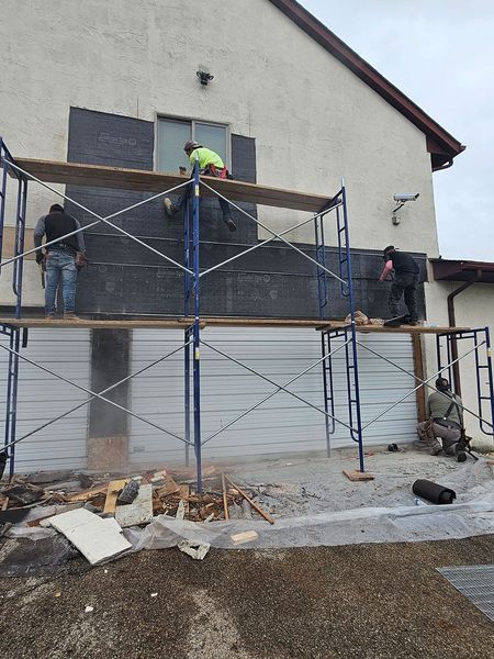 Four workers on scaffolding repair a house exterior, installing dark, waterproof building paper over the white stucco wall.