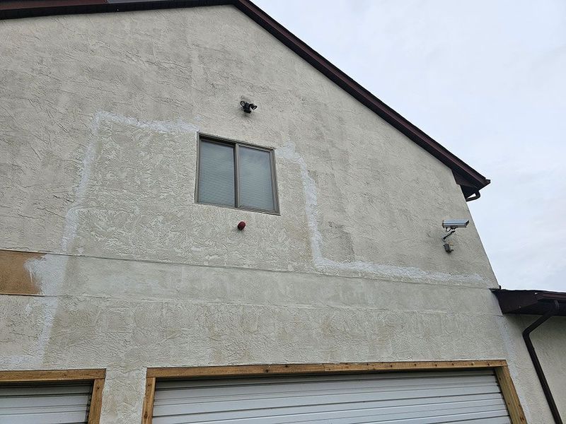 Exterior stucco wall of a house with a window, security camera, and light, showing signs of patch repair around the window.