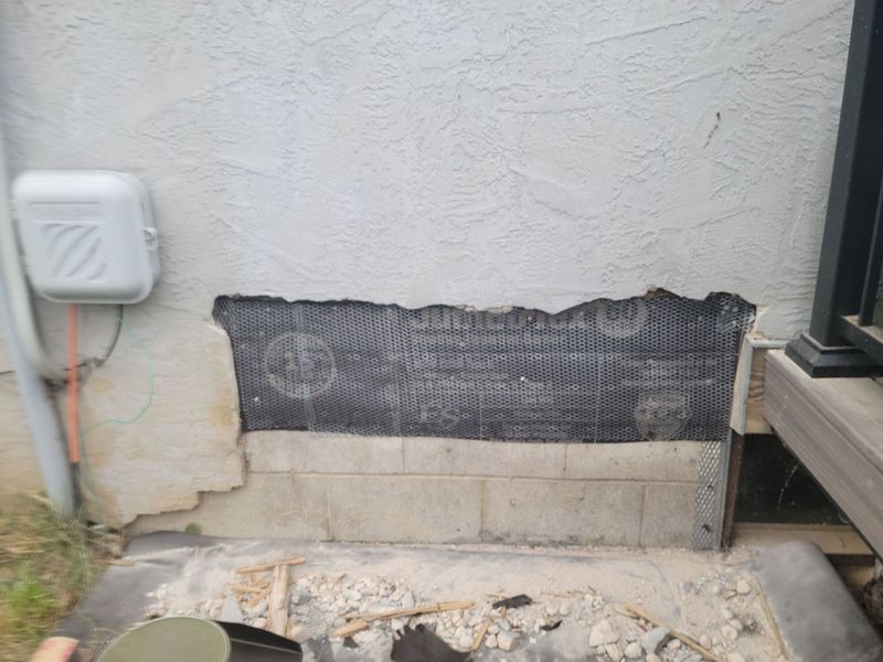 A damaged exterior stucco wall revealing a black moisture barrier and concrete blocks next to a deck and utility box.