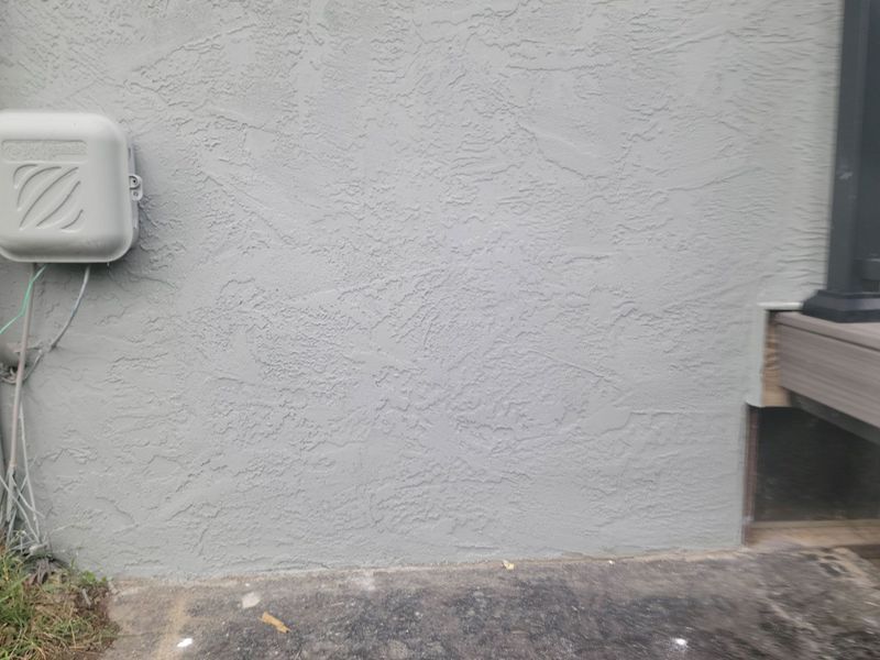 A light gray textured exterior stucco wall with a utility box on the left and a deck support on the right.