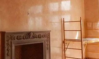 An ornate fireplace mantel sits before a peach-colored wall with a tall, metal scaffolding unit beside it.