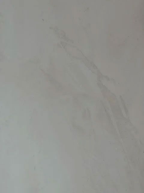 A close-up of a plain, light grey wall with subtle, diagonal textural streaks and slight variations in shade.