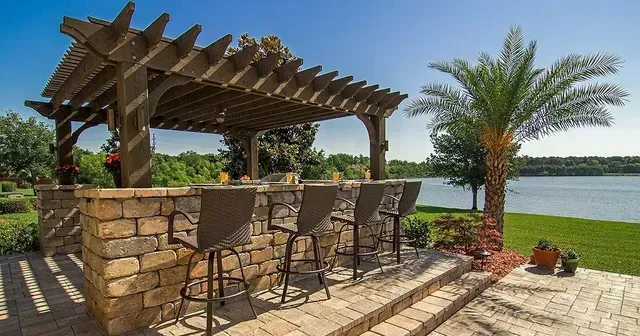 Outdoor bar with stone facade, wooden pergola, and seating overlooking a lake.