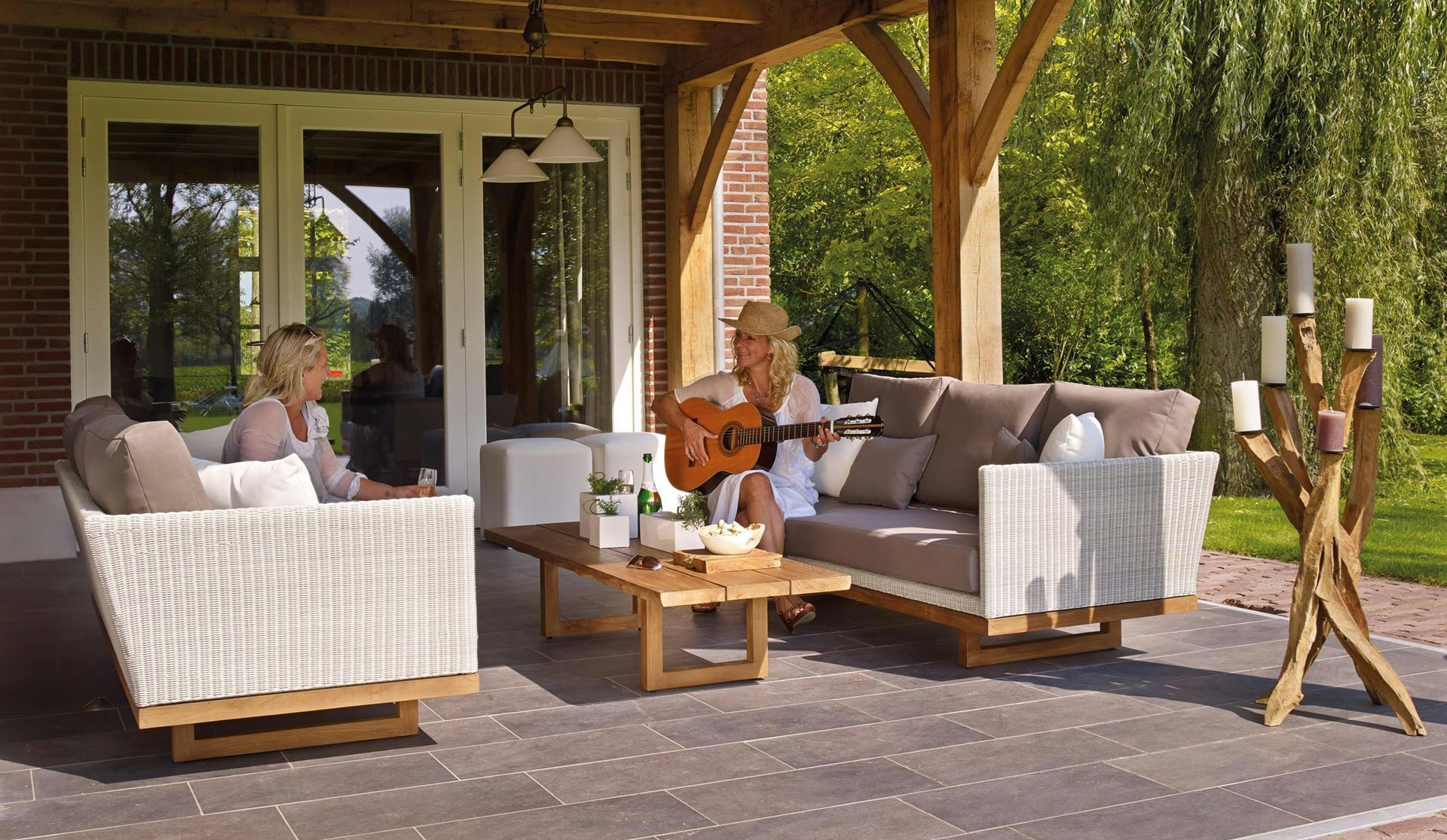 Two people on patio furniture, one playing guitar. Outdoor setting with a wooden pergola.