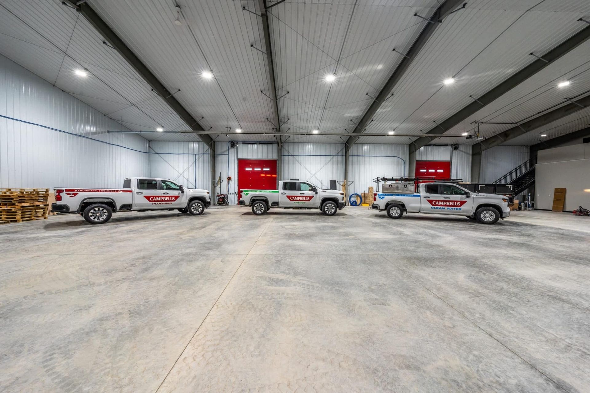 Three white company trucks parked inside a large warehouse with red doors.
