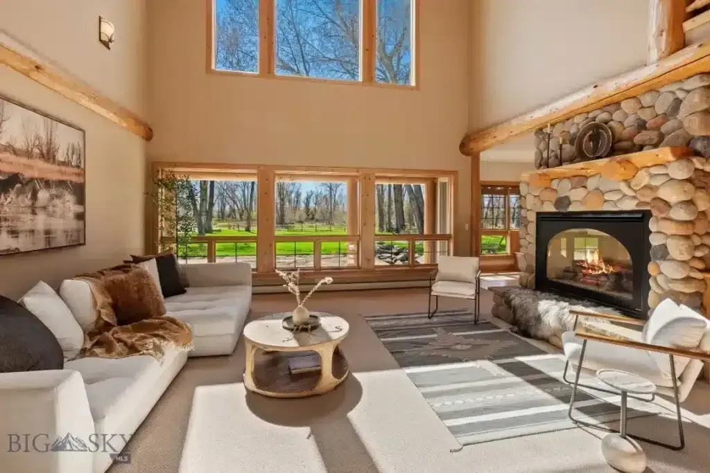 Living room with stone fireplace, large windows overlooking green space, white sofas, and neutral color scheme.