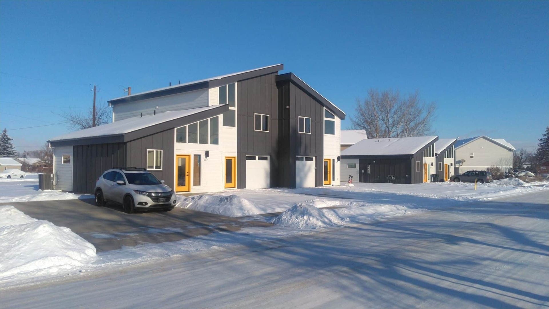 Modern two-story building with a silver SUV parked in the snowy driveway. Gray and white exterior with yellow trim.