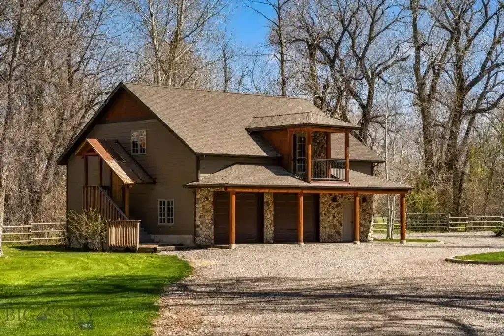 Two-story brown house with stone accents, carport, and porch, set in a wooded area with gravel driveway.