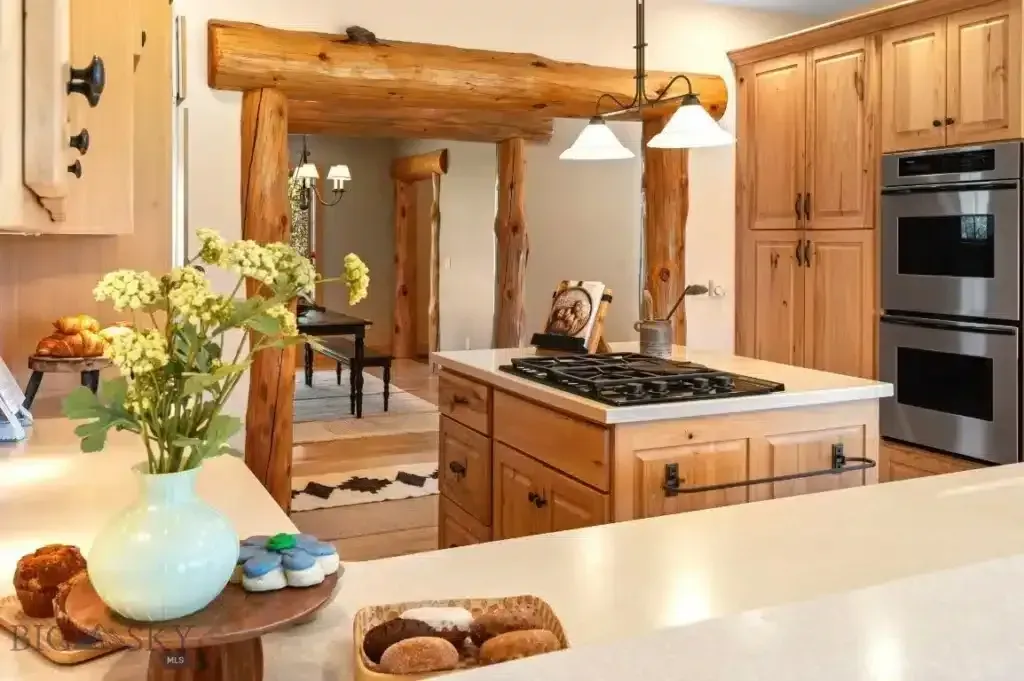 Kitchen with light wood cabinets, island with stovetop, and a log-framed doorway.