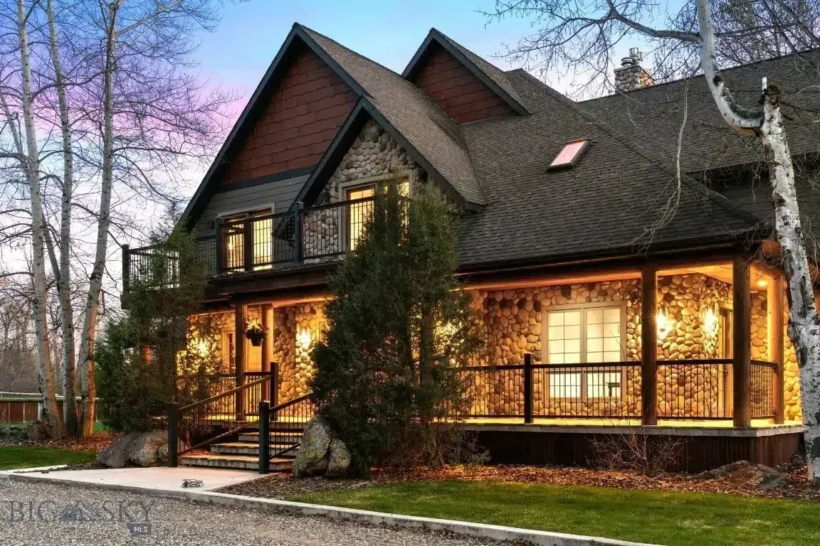 Stone and wood house with a wraparound porch, lit from within, under a dusk sky.
