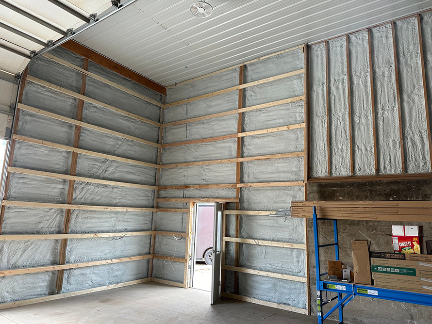 Interior view of a building with wooden framing and spray foam insulation. A door and garage door are visible.