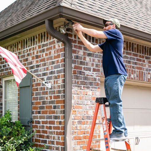 A man is standing on a ladder fixing a light on the side of a brick house.