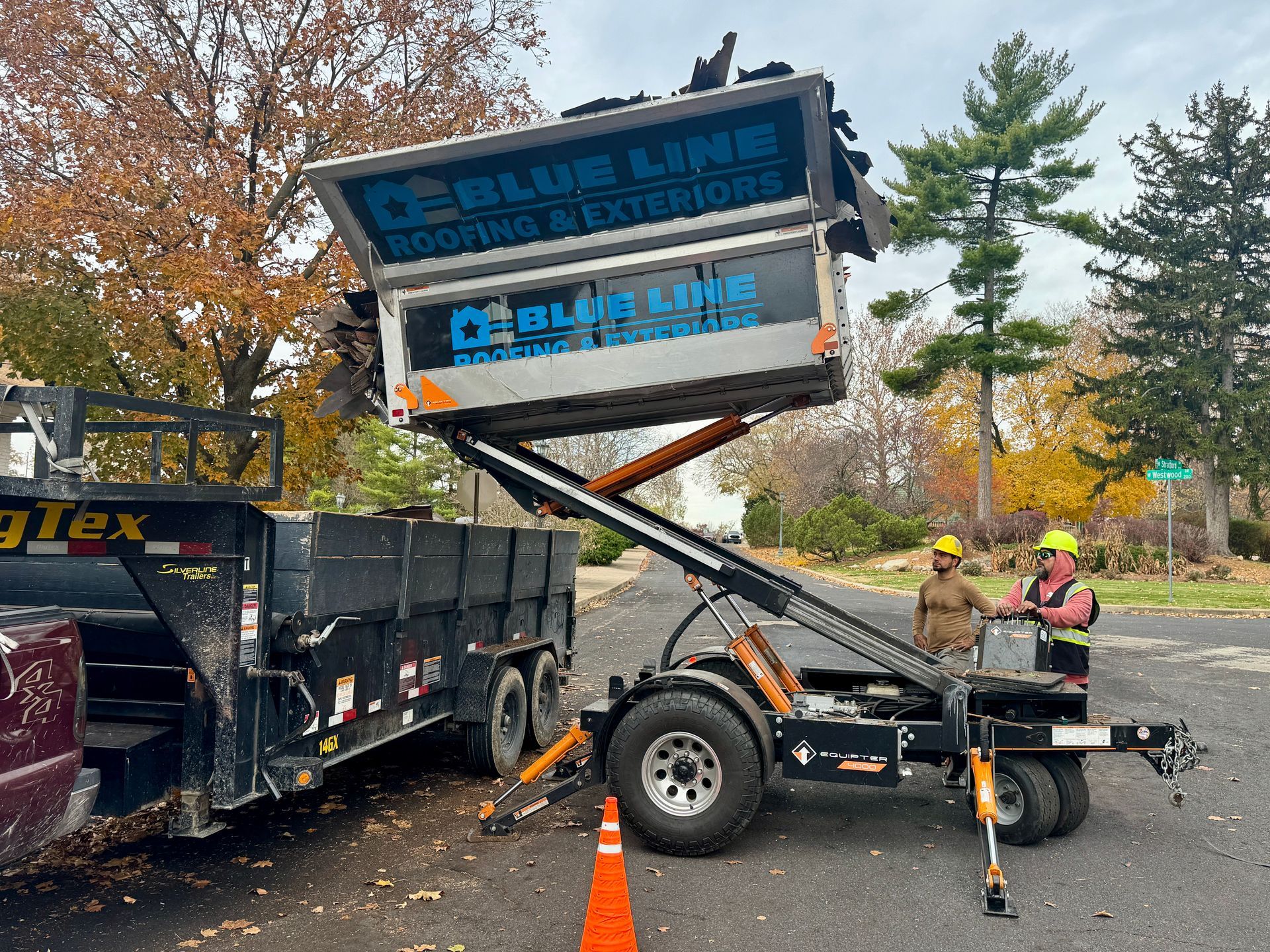 Roofing debris being loaded into a trailer with an elevated lift; two workers in vests.