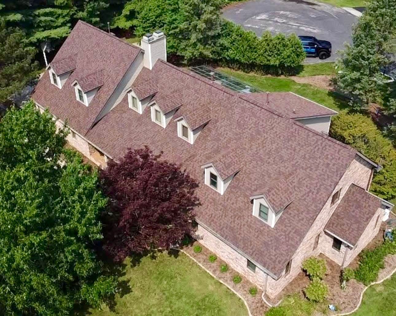 Brown-roofed house with dormers, brick exterior, and surrounding greenery.