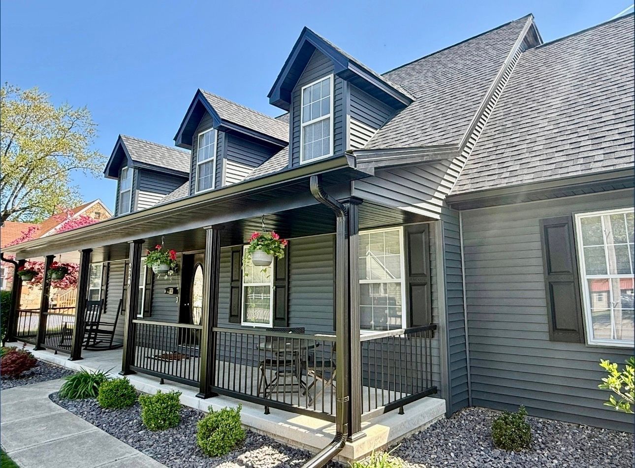 Dark gray house with a porch, black trim, and three dormers. Flowers hang from the porch.