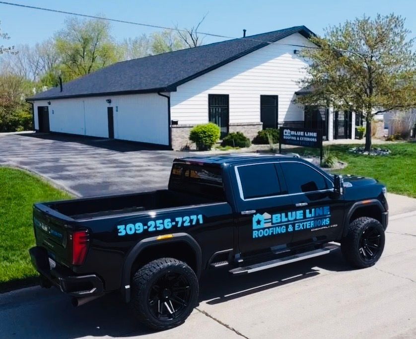 Black truck with Blue Line Roofing & Exteriors logo parked in front of a white building with black roof.