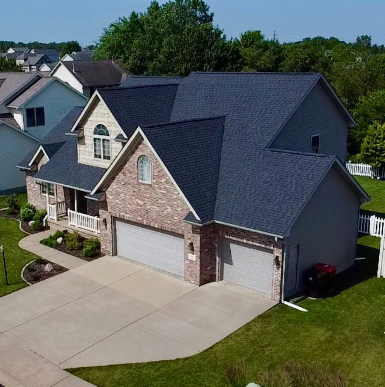 Two-story brick house with dark blue roof, two-car garage, and concrete driveway on a sunny day.