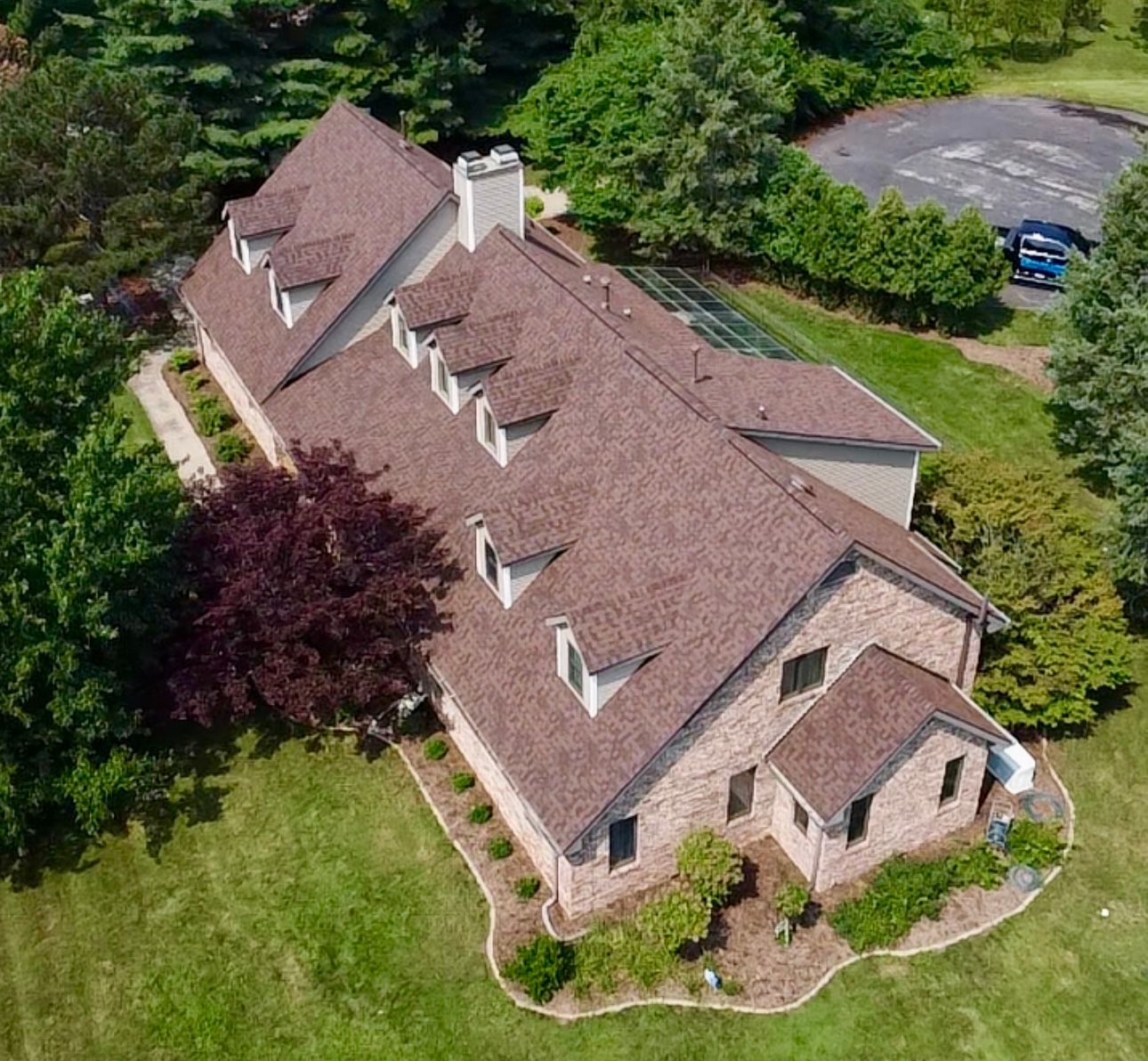 Brown-roofed house with multiple dormers, brick facade, surrounded by green grass and trees.