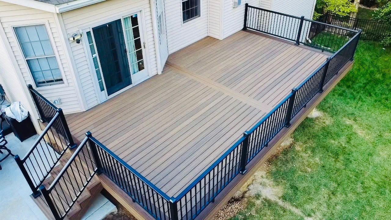 Elevated deck with composite wood boards, black railing, and stairs, next to a two-story beige house on a grassy lawn.
