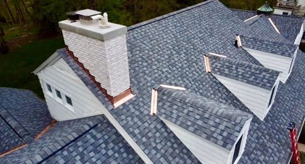 An aerial view of a gray shingled roof with a white brick chimney and multiple dormer windows.