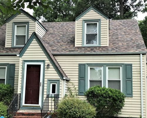 Beige house with a brown roof, teal shutters and trim, and a dark red door.