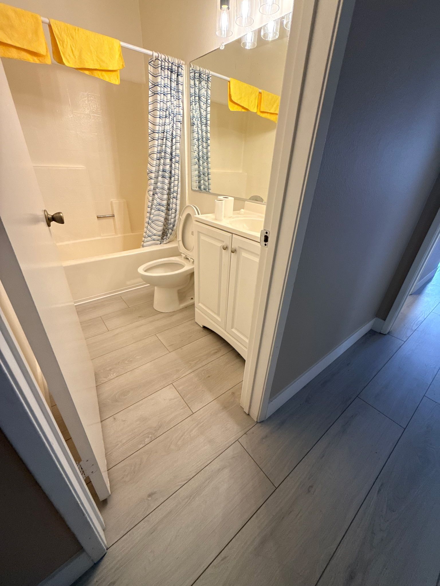A bathroom with grey wood-look flooring, a white vanity, a toilet, a bathtub with a patterned curtain, and yellow towels.