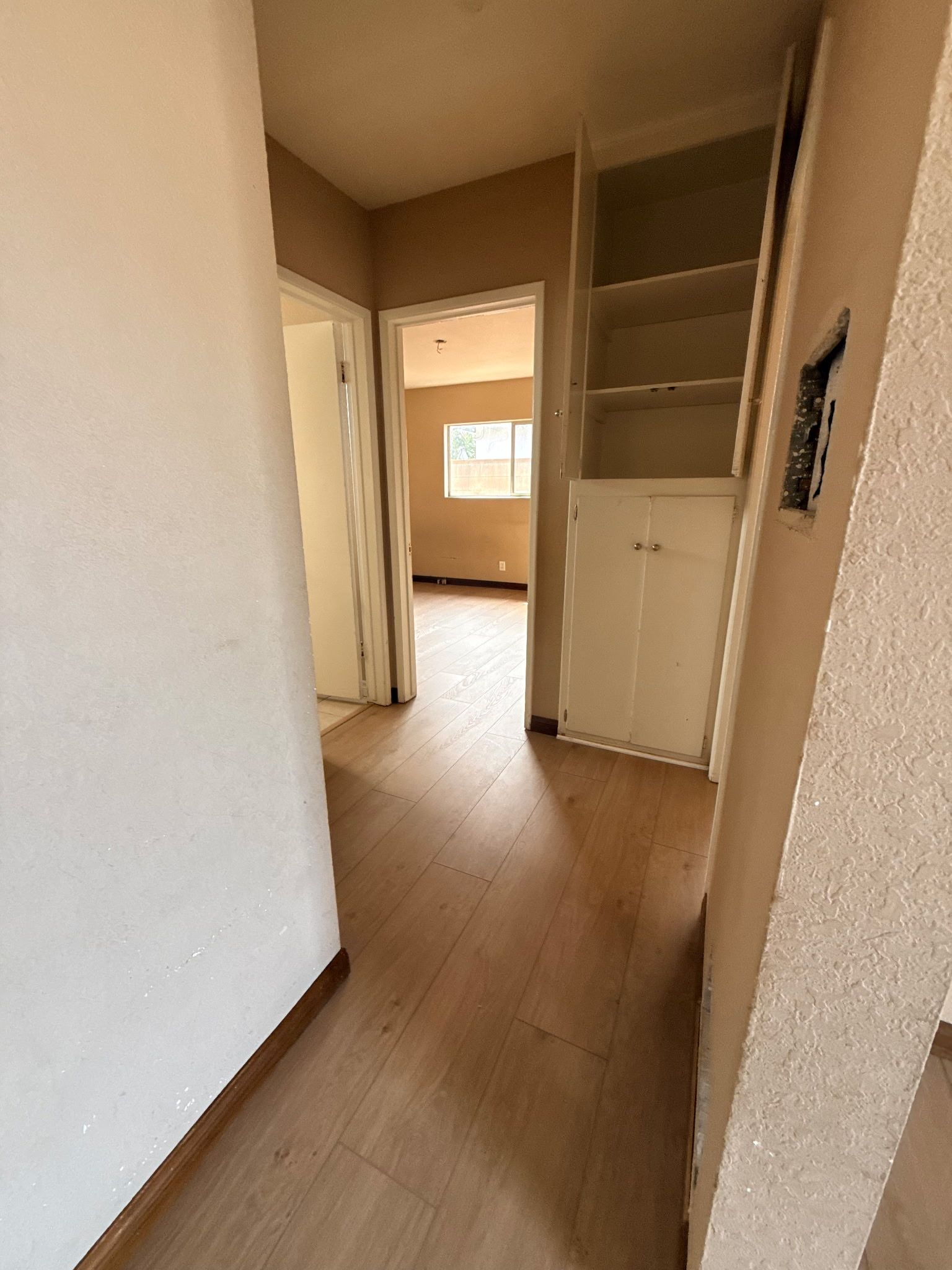 A hallway with light wood-style flooring leads to a doorway showing a room with a window, alongside a white built-in shelf.