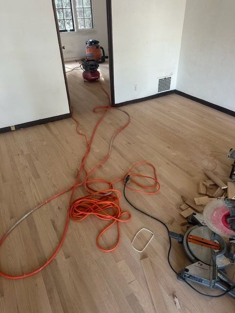 Interior room with freshly sanded hardwood floors, a miter saw on the right, and vacuum equipment in the adjacent doorway.