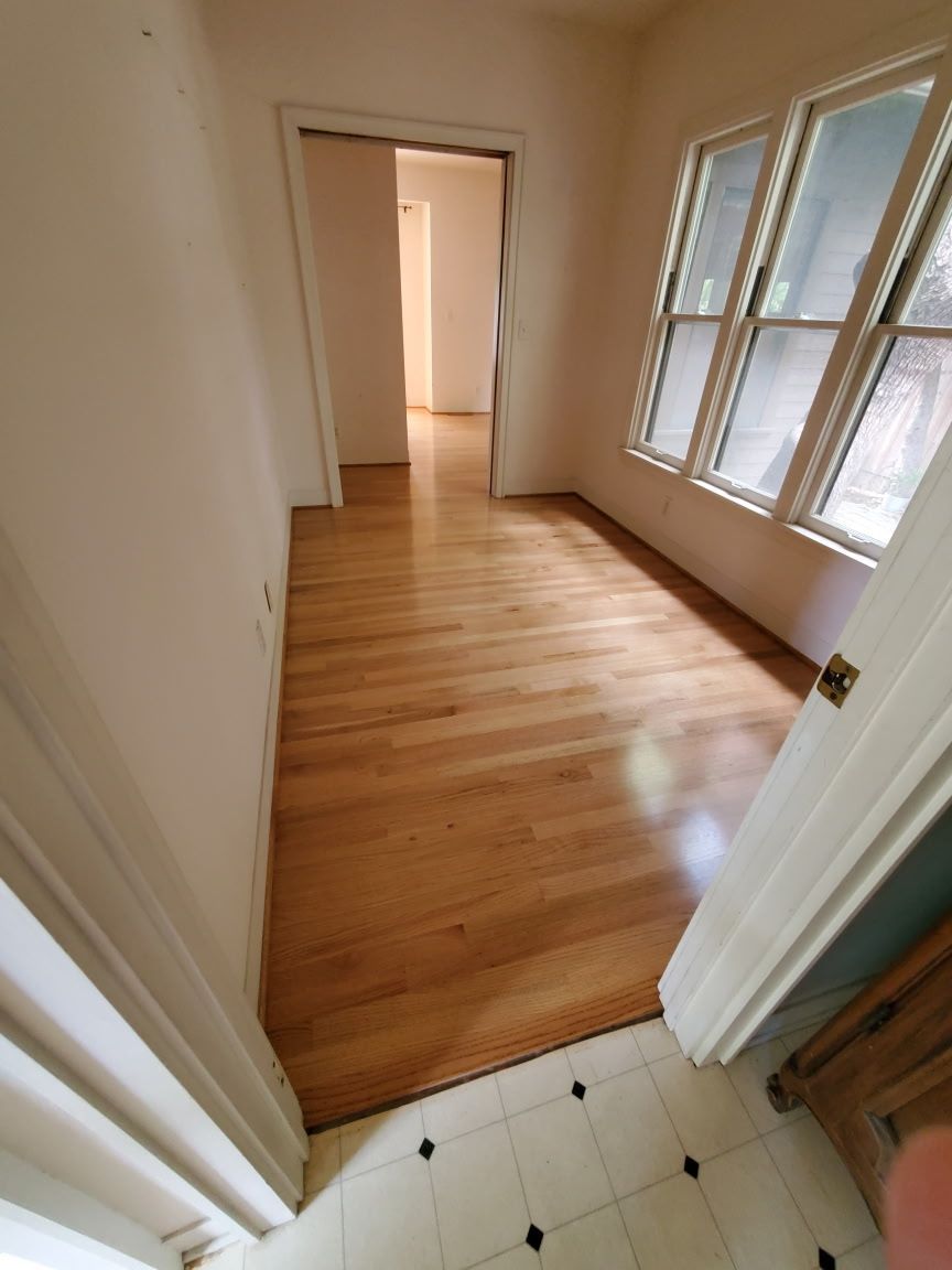 An interior view from a tiled entryway looking into a sunlit room with polished wood floors and a doorway to another room.