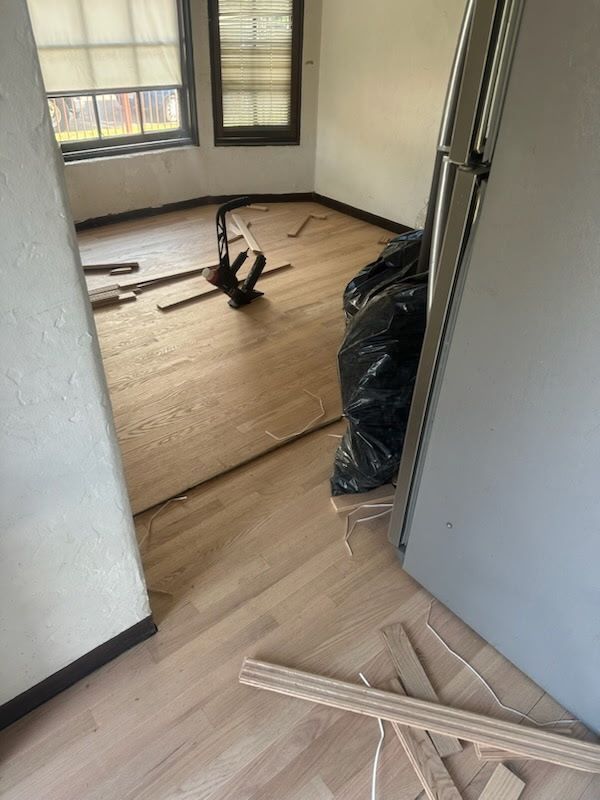 A room with newly installed light wood flooring, floor trim pieces, and a black trash bag near an open doorway.