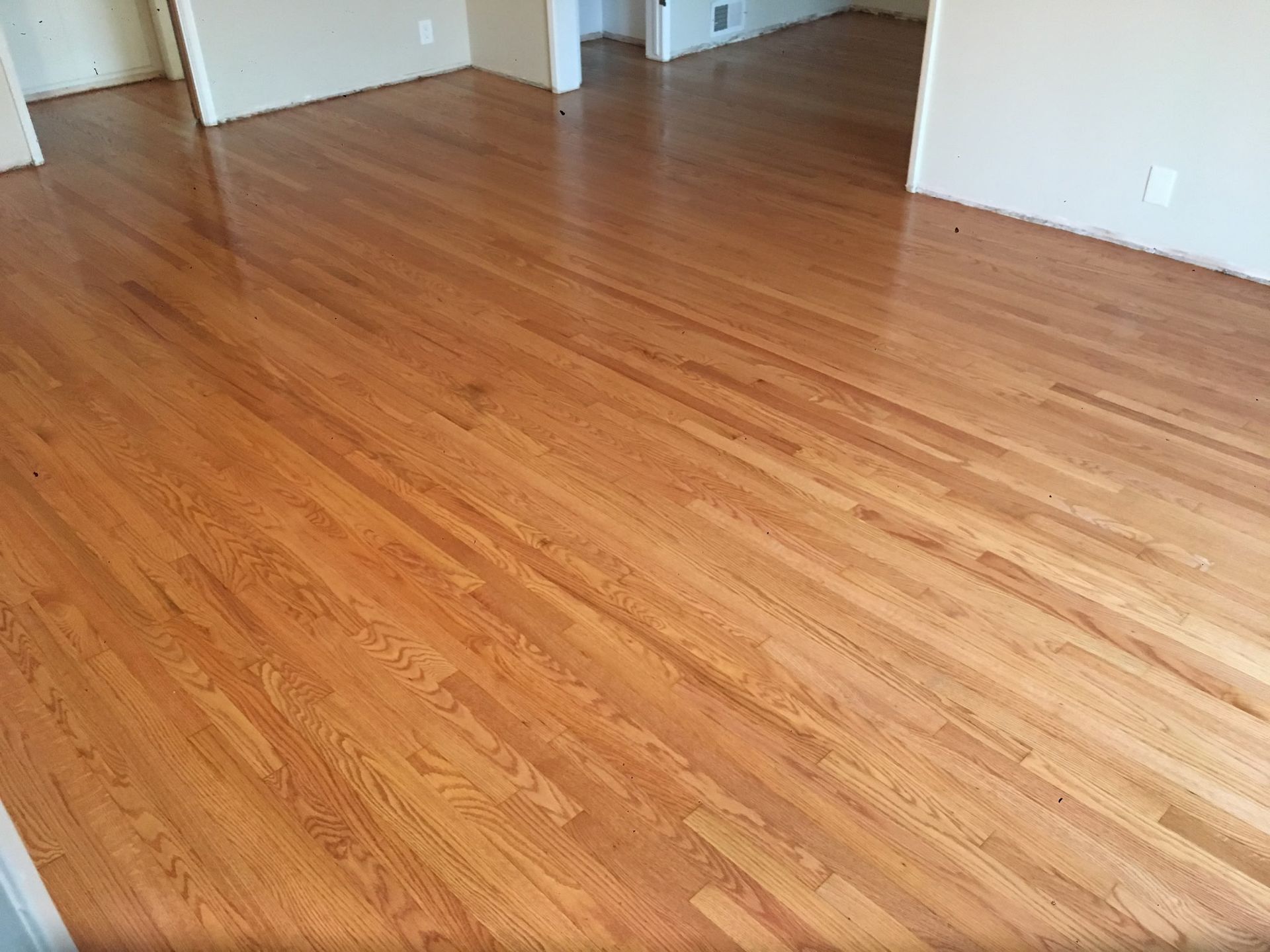 A room featuring polished light oak hardwood flooring with visible wood grain, white walls, and open doorways.
