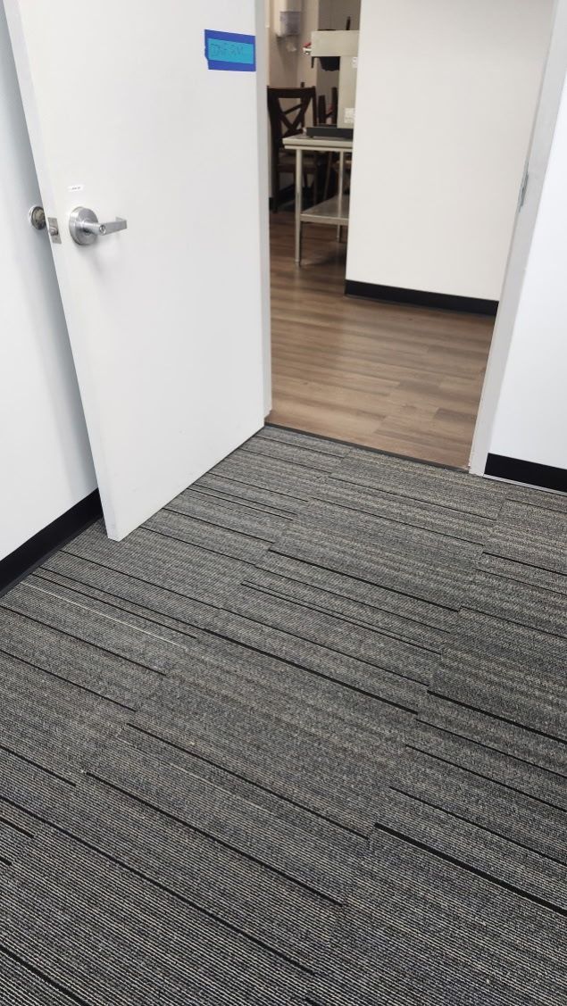 Open white door leading from a gray carpeted office into a room with wood flooring and a small table.