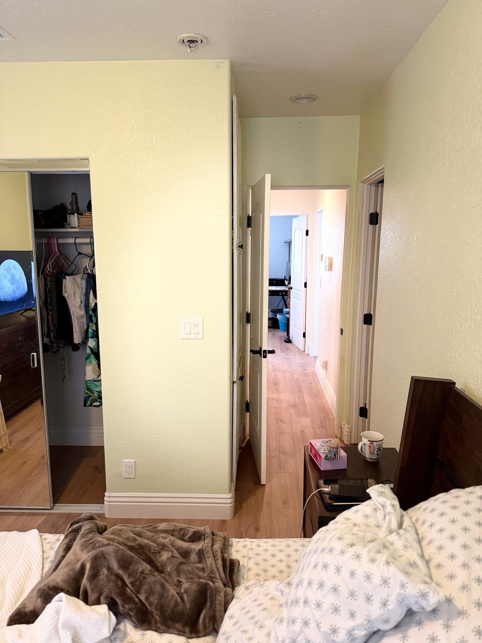 A bedroom with light yellow walls, a closet on the left, and a hallway leading to another room on the right.