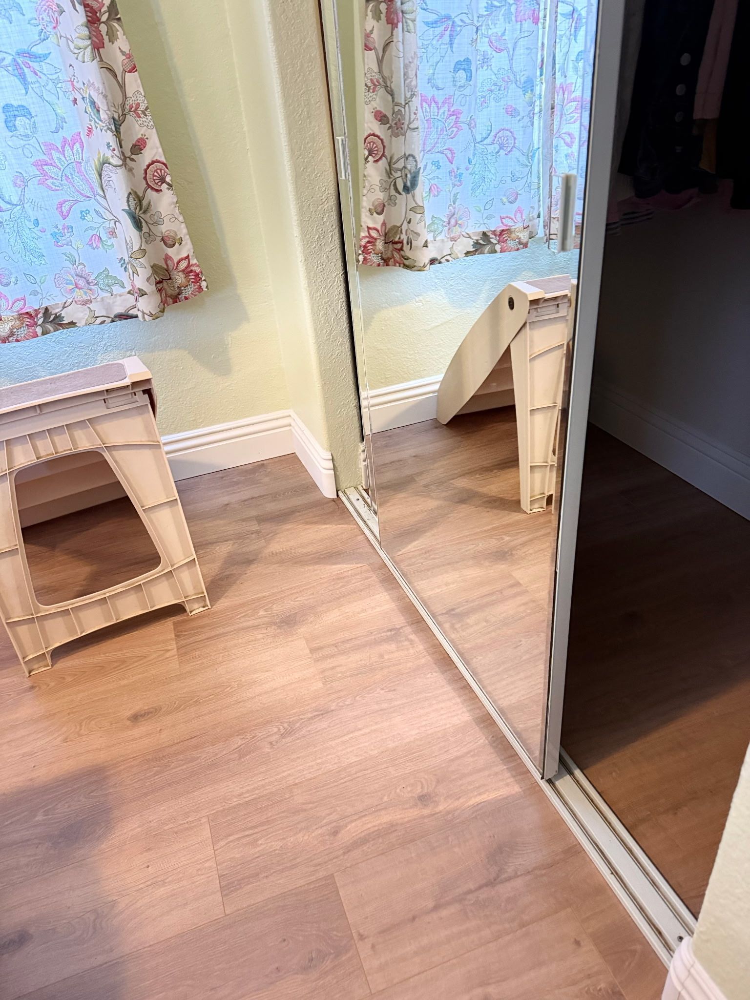 A light wood floor meets a mirrored closet door, reflecting a white stool and floral curtains in a bright room.