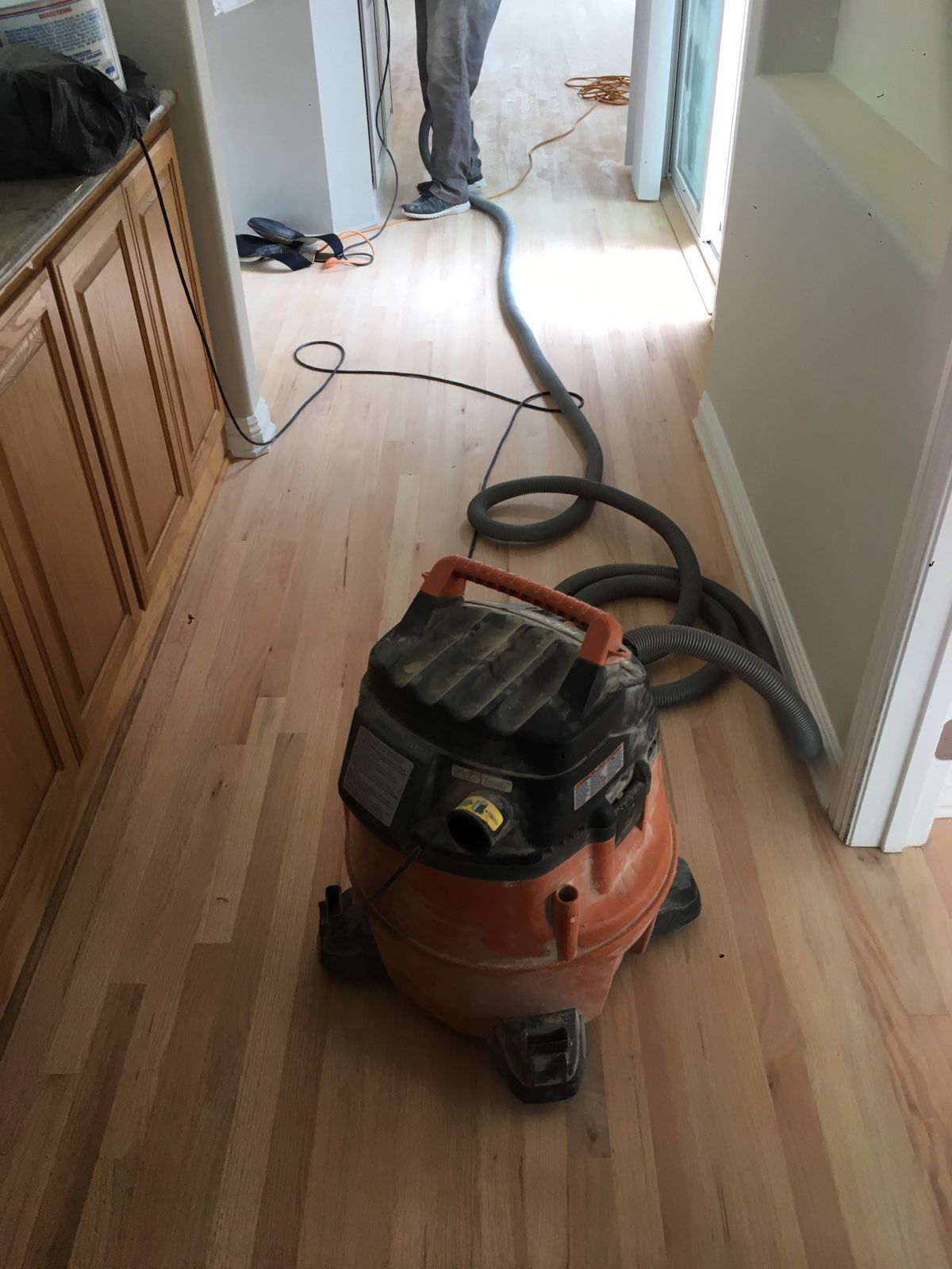 An orange shop vacuum with a hose extending across a freshly sanded hardwood floor in a home.